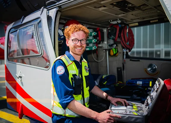 Lächelnder männlicher Sanitäter mit roten Haaren und Brille, der neben einem Hubschrauber steht und sich um die medizinische Notfallausrüstung im Flugzeug kümmert.