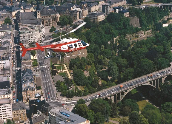 Rot-weißer Hubschrauber, der über einen Stadtpark mit einer Brücke und Straßen fliegt, die von Gebäuden und Bäumen umgeben sind.