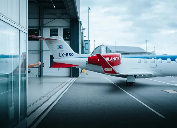 Vue latérale arrière d'un avion d'ambulance médicale blanc et rouge immatriculé LX-RSQ stationné sur le tarmac de l'aéroport près d'un hangar avec le bras d'une personne tendu par une porte vitrée.