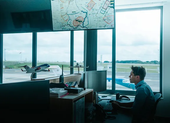 Homme portant un casque assis à un bureau avec plusieurs écrans dans la salle de contrôle d'un aéroport, regardant par la fenêtre un avion en stationnement.