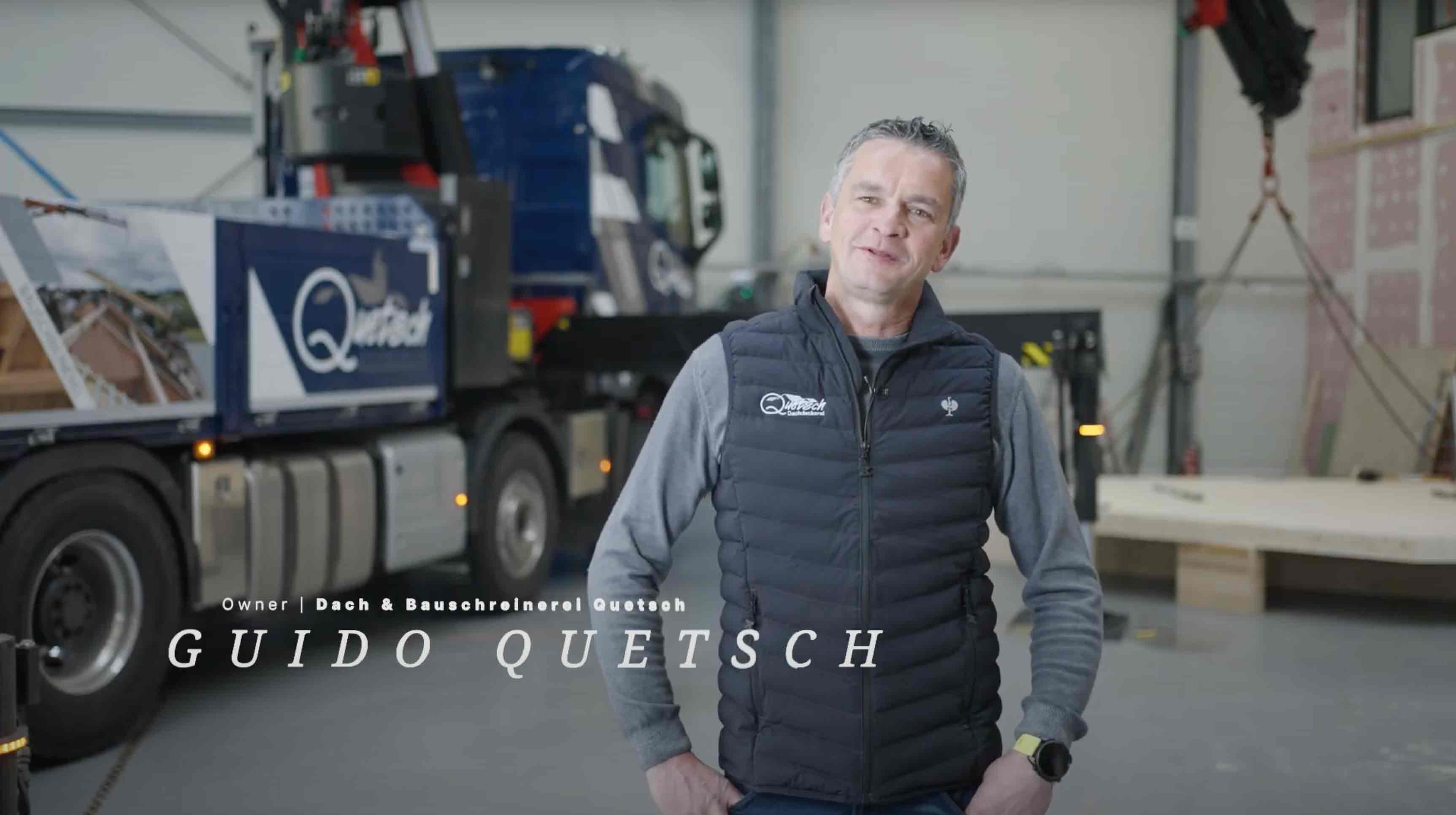 Mann in schwarzer Weste mit Firmenlogos steht in einem Lagerhaus mit einem Marken-Truck im Hintergrund.