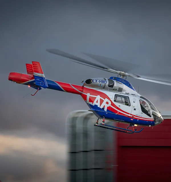 Red, white, and blue helicopter with 'LAR' logo flying against a cloudy sky near a building.