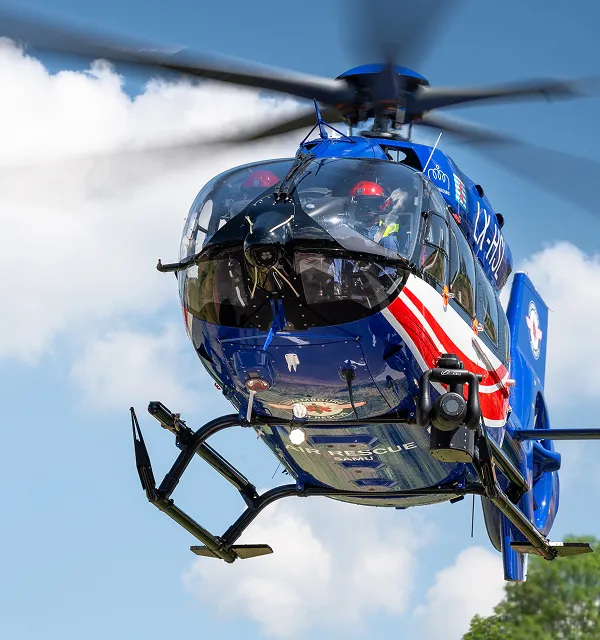 Blue air rescue helicopter flying with two crew members visible inside the cockpit against a partly cloudy sky.