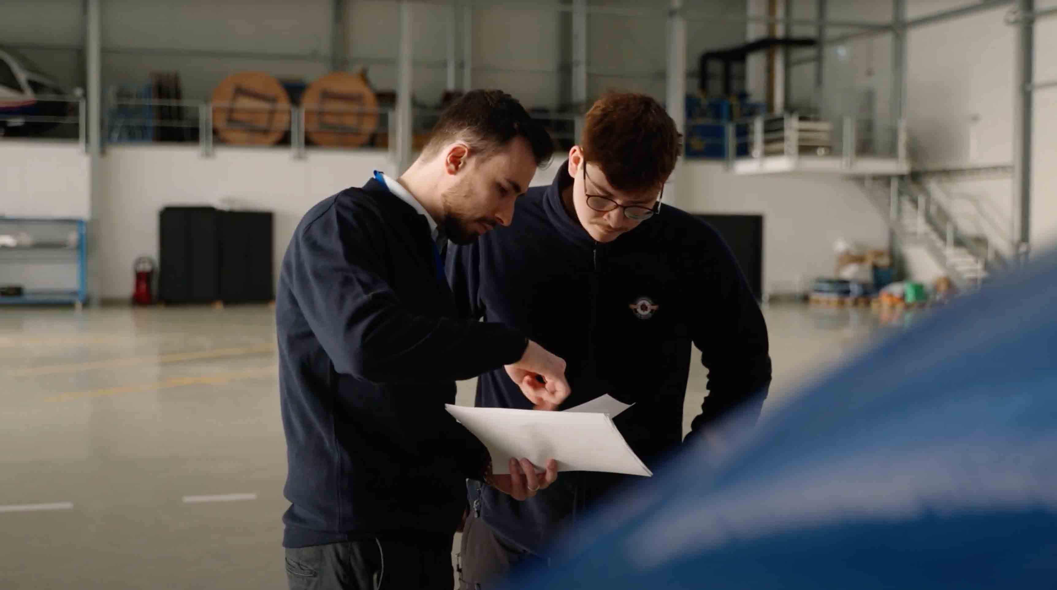 Two men indoors looking at documents, with industrial equipment and storage visible in the background.