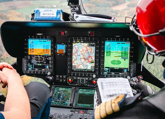 View of a helicopter cockpit with digital navigation and control displays, map screen, and pilots operating the controls and holding a checklist.
