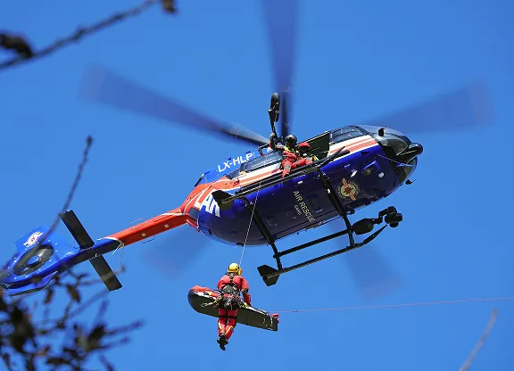 Air rescue helicopter with crew member being lowered on a stretcher during a rescue operation against clear blue sky.
