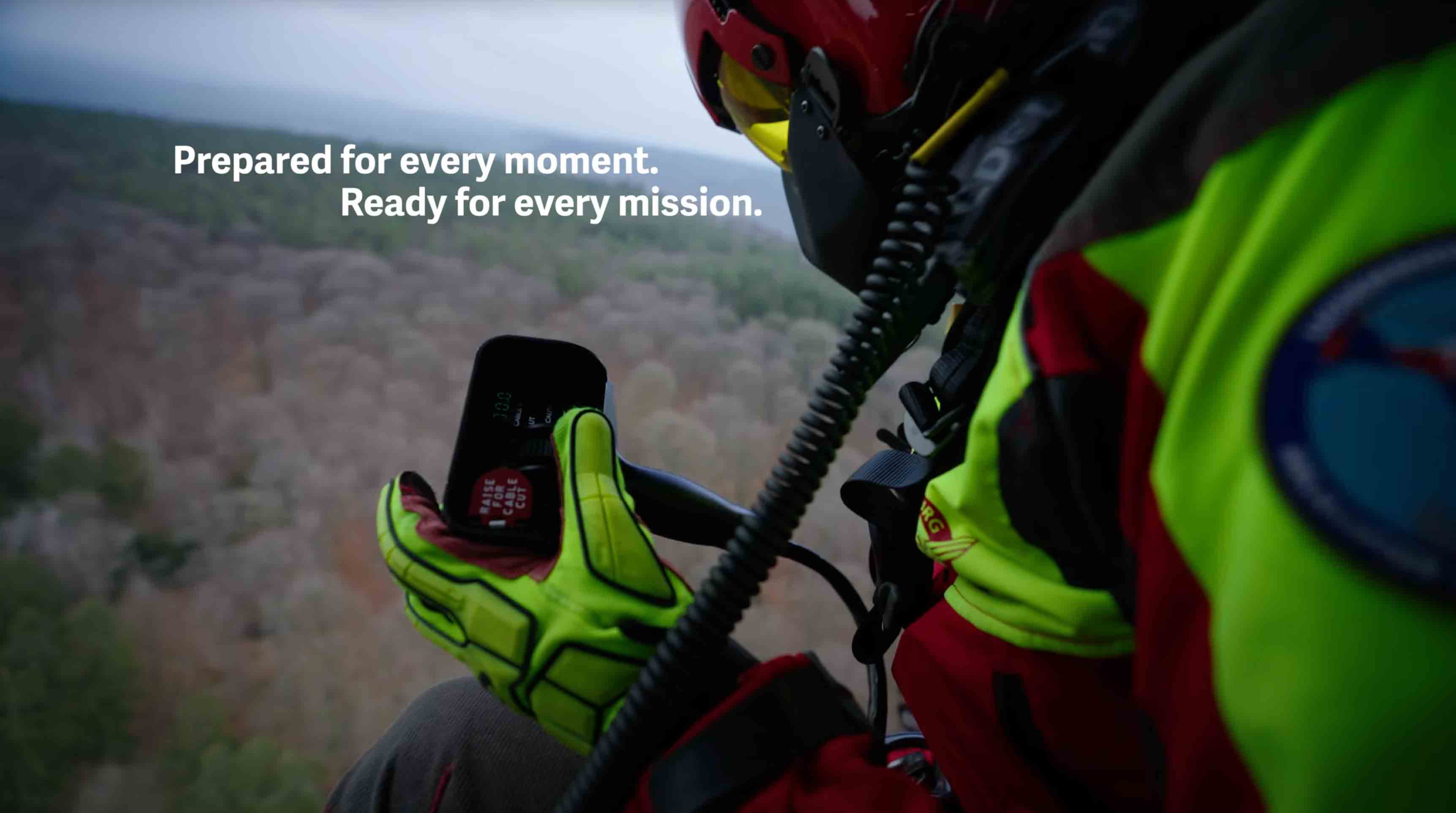 Rescue worker wearing a bright yellow glove and helmet operates a radio while suspended above a forest during a helicopter mission.