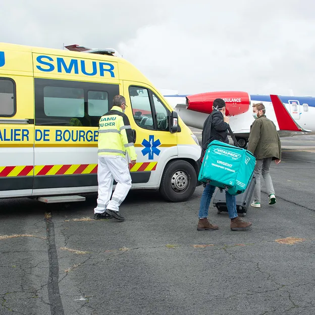 Medical personnel near a yellow SMUR ambulance on a tarmac, with two men carrying urgent transport medical bags walking towards an aircraft marked Ambulance.