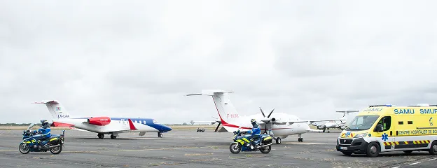 Two motorcyclists in blue and yellow uniforms riding on an airport tarmac near a small white and blue jet, a white propeller plane, and a yellow SAMU ambulance.