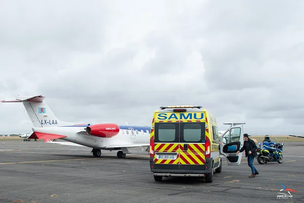 Emergency medical ambulance van with SAMU markings parked on an airstrip near a small white and red private jet, with a person walking towards the vehicle and a police motorcycle in the background under a cloudy sky.