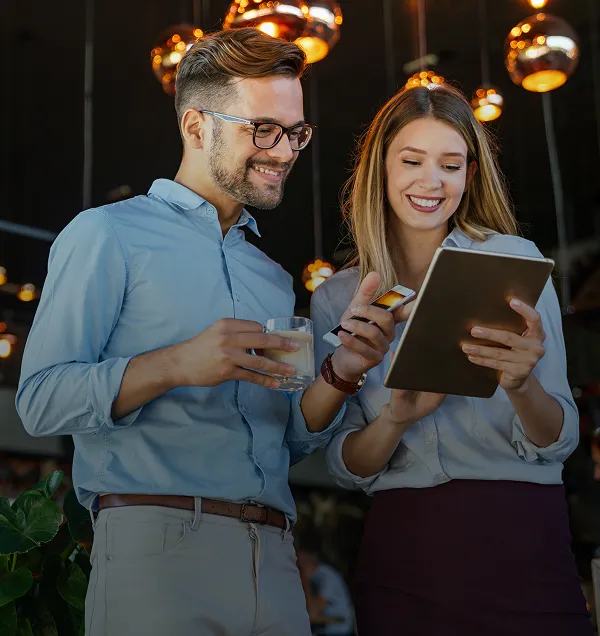 Mann und Frau lächeln, während sie in einer warm beleuchteten Umgebung auf ein digitales Tablet schauen.