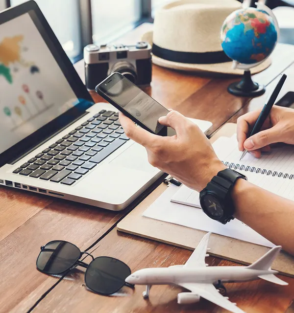 Person, die ein Smartphone benutzt und in ein Notizbuch an einem Holzschreibtisch mit Laptop, Spielzeugflugzeug, Sonnenbrille, Kamera und Globus schreibt.