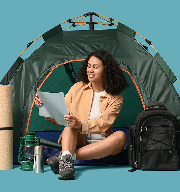 Lächelnde Frau sitzt mit gekreuzten Beinen vor einem grünen Campingzelt und liest eine Zeitung. In der Nähe befinden sich eine gerollte Schlafmatte, eine Laterne, eine Wasserflasche und ein schwarzer Rucksack.