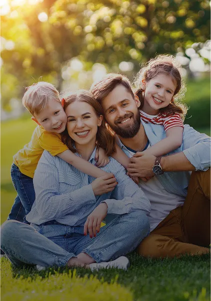 Lächelnde vierköpfige Familie sitzt auf Gras in einem sonnenbeschienenen Park, Eltern sitzen mit Kindern, die sie umarmen.
