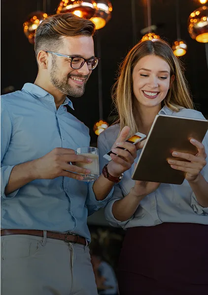 Lächelnder Mann und Frau schauen auf ein Tablet, auf dem der Mann ein Glas Wasser in einem warm beleuchteten Raum hält.