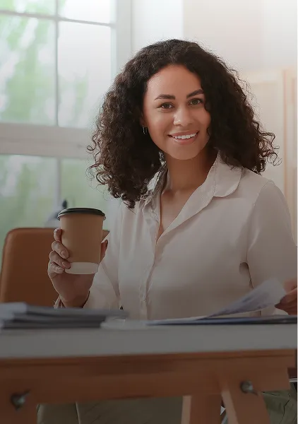 Lächelnde Frau mit lockigem Haar, die eine Kaffeetasse zum Mitnehmen und einige Zeitungen in der Hand hält, während sie an einem Schreibtisch sitzt.