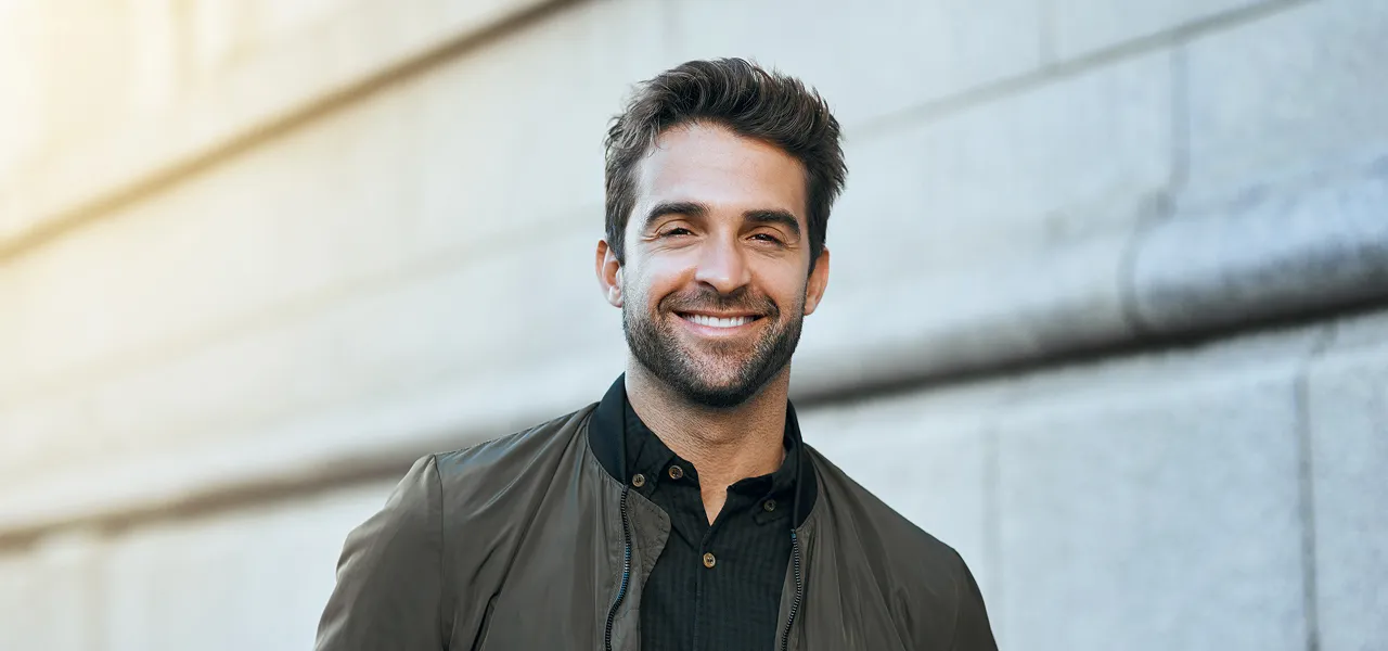 Lächelnder Mann mit kurzen dunklen Haaren und Bart in dunkler Jacke und Hemd, der draußen in der Nähe einer grauen Steinmauer steht.