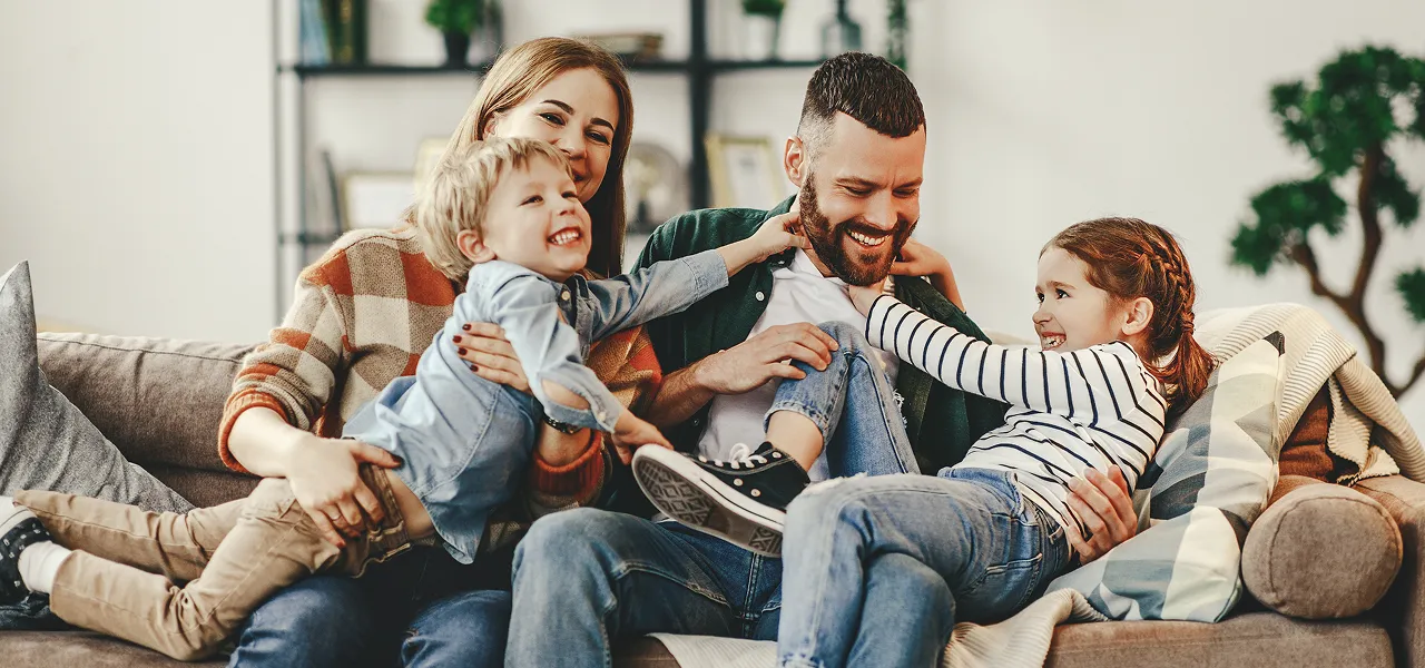 Happy family of four playing and laughing together on a living room sofa.