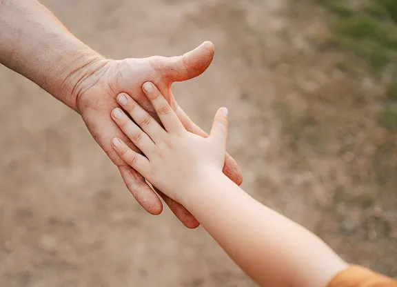 Adult hand and child's hand reaching to touch each other outdoors on a dirt path.