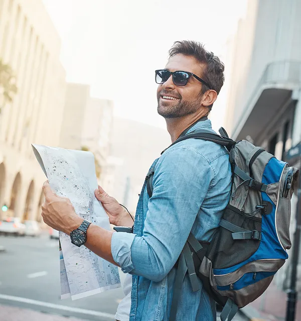 Lächelnder Mann mit Sonnenbrille und blauem Jeanshemd, der einen Stadtplan mit einem großen Rucksack auf dem Rücken in einer urbanen Straßenumgebung hält.