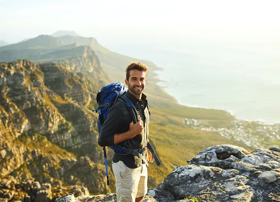 Lächelnder Mann mit Rucksack, der auf einem felsigen Berg mit Blick auf das Tal und das ferne Meer steht.