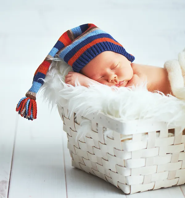 Nouveau-né endormi portant un bonnet en tricot rayé, allongé sur une couverture blanche moelleuse à l'intérieur d'un panier tissé blanc.