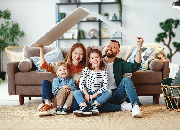 Happy family of four sitting on floor in living room, parents holding a cardboard roof over children.