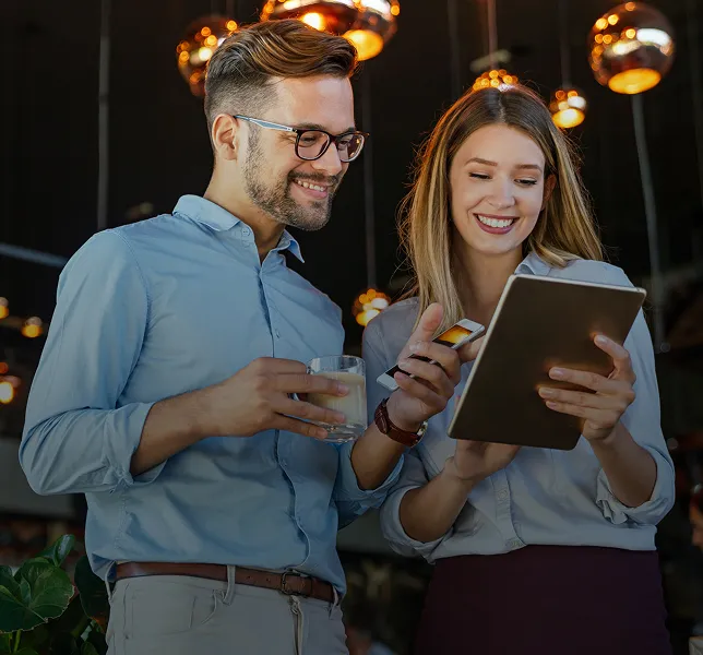 Lächelnder Mann und Frau schauen gemeinsam in einem warm beleuchteten Raum auf eine Tablette, wobei der Mann ein Glas Wasser hält und die Frau eine Tablette in der Hand.