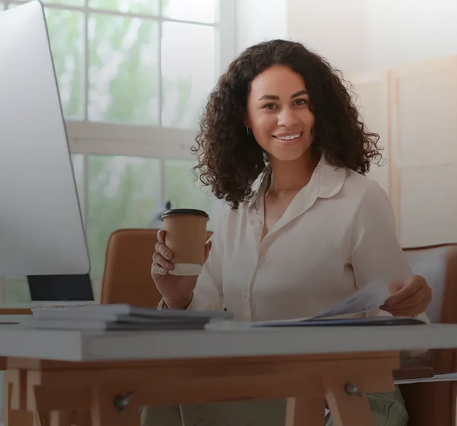 Frau mit lockigem Haar sitzt an einem Schreibtisch, hält eine Kaffeetasse und Papiere in der Hand und lächelt in die Kamera.