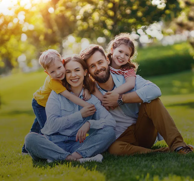 Glückliche vierköpfige Familie, die draußen auf Gras sitzt, Kinder umarmen ihre Eltern und lächeln.