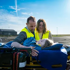 Deux parents vêtus de gilets jaunes à haute visibilité rassurent leur enfant allongé sur une civière à l'extérieur sous un ciel bleu.