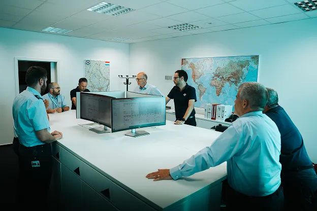 Groupe de sept hommes dans une salle de réunion autour d'une grande table blanche avec plusieurs écrans d'ordinateur et des cartes du monde accrochées aux murs.