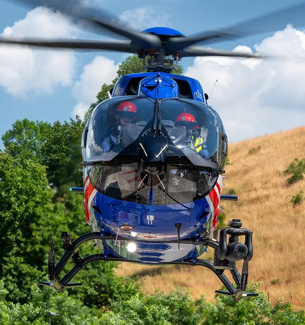 Blue rescue helicopter flying with two crew members wearing red helmets visible through the cockpit window against a background of trees and a grassy hill.