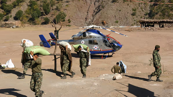 Soldiers carrying large sacks of supplies in a dry, mountainous area with a parked grey and blue helicopter in the background.