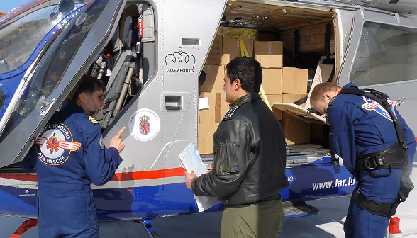 Three rescue workers beside a helicopter loaded with cardboard boxes, one wearing a Luxembourg Air Rescue uniform.