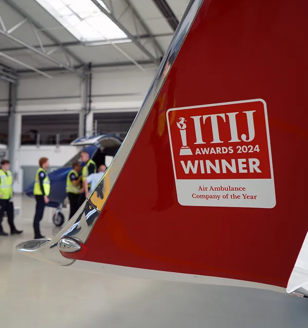 Nageoire arrière rouge d'un avion avec un autocollant indiquant la compagnie d'ambulance aérienne gagnante des ITIJ Awards 2024, avec des personnes et un avion bleu en arrière-plan à l'intérieur d'un hangar.