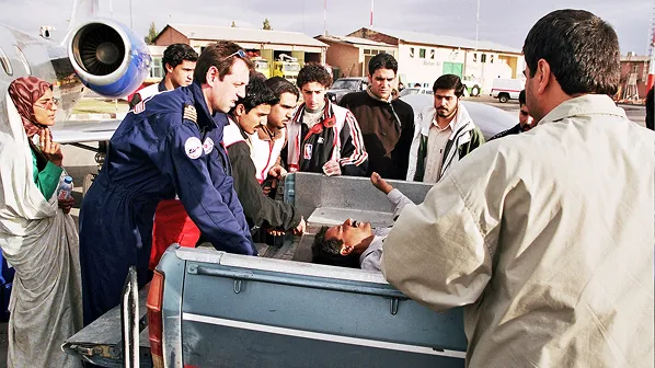 A group of people surrounding a man lying in the back of a blue pickup truck near an airplane on a tarmac.