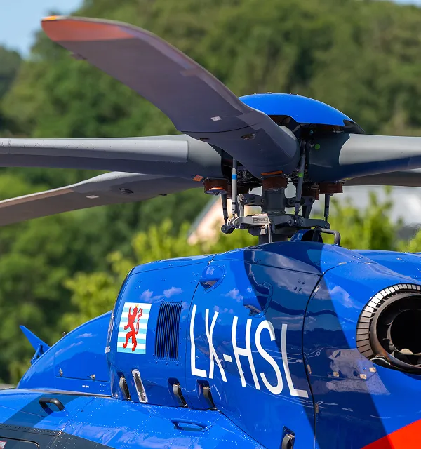 Blue helicopter with registration LX-HSL and a red lion emblem on the side, parked outdoors with trees in the background.
