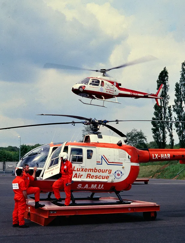 Two Luxembourg Air Rescue helicopters with red and white livery, one on the ground with three crew members in red uniforms and one flying in the sky.
