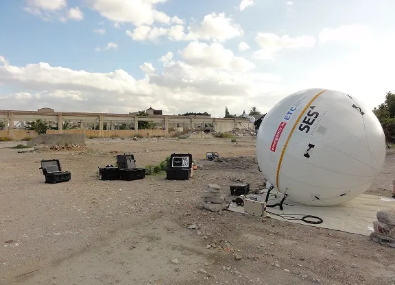 Un gros ballon blanc de communication d'urgence SES est installé sur une bâche au sol dans une zone aride avec des boîtiers d'équipement éparpillés à proximité et des bâtiments partiellement endommagés en arrière-plan sous un ciel nuageux.