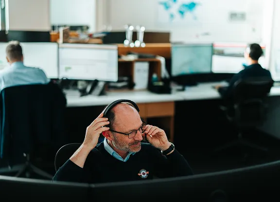 Un homme d'âge moyen portant des lunettes met un casque dans un bureau avec plusieurs écrans d'ordinateur et des collègues travaillant en arrière-plan.