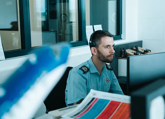 Agent de sécurité portant un casque et un uniforme bleu assis à un bureau avec des écrans d'ordinateur dans un bureau.