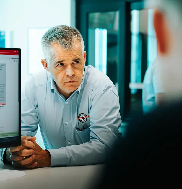 Homme sérieux d'âge moyen vêtu d'une chemise boutonnée bleu clair parlant à quelqu'un de l'autre côté d'un bureau avec un écran au premier plan.