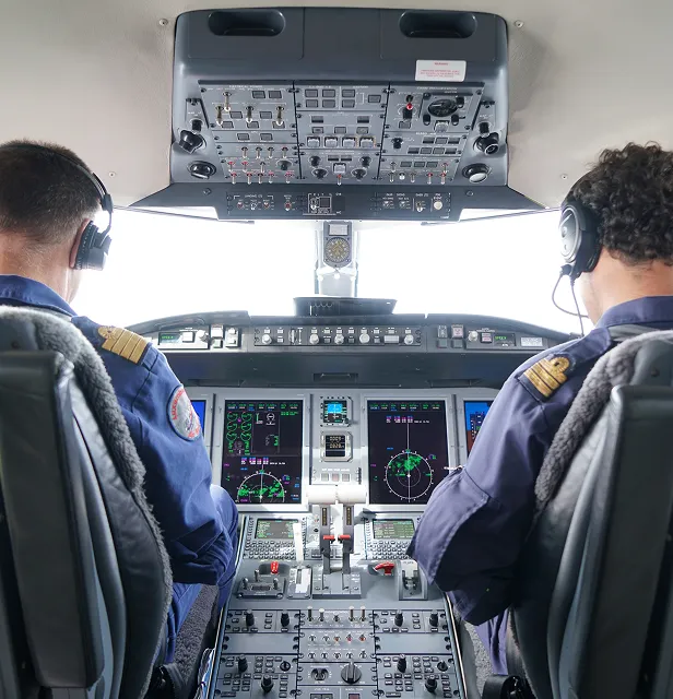 Deux pilotes portant des casques sont assis dans un poste de pilotage d'avion moderne doté de plusieurs panneaux de commande et d'écrans numériques.