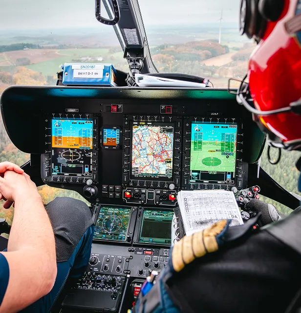 Poste de pilotage d'hélicoptère avec deux pilotes, montrant des instruments de vol, des écrans de navigation et un pilote portant un casque rouge tenant une liste de contrôle.