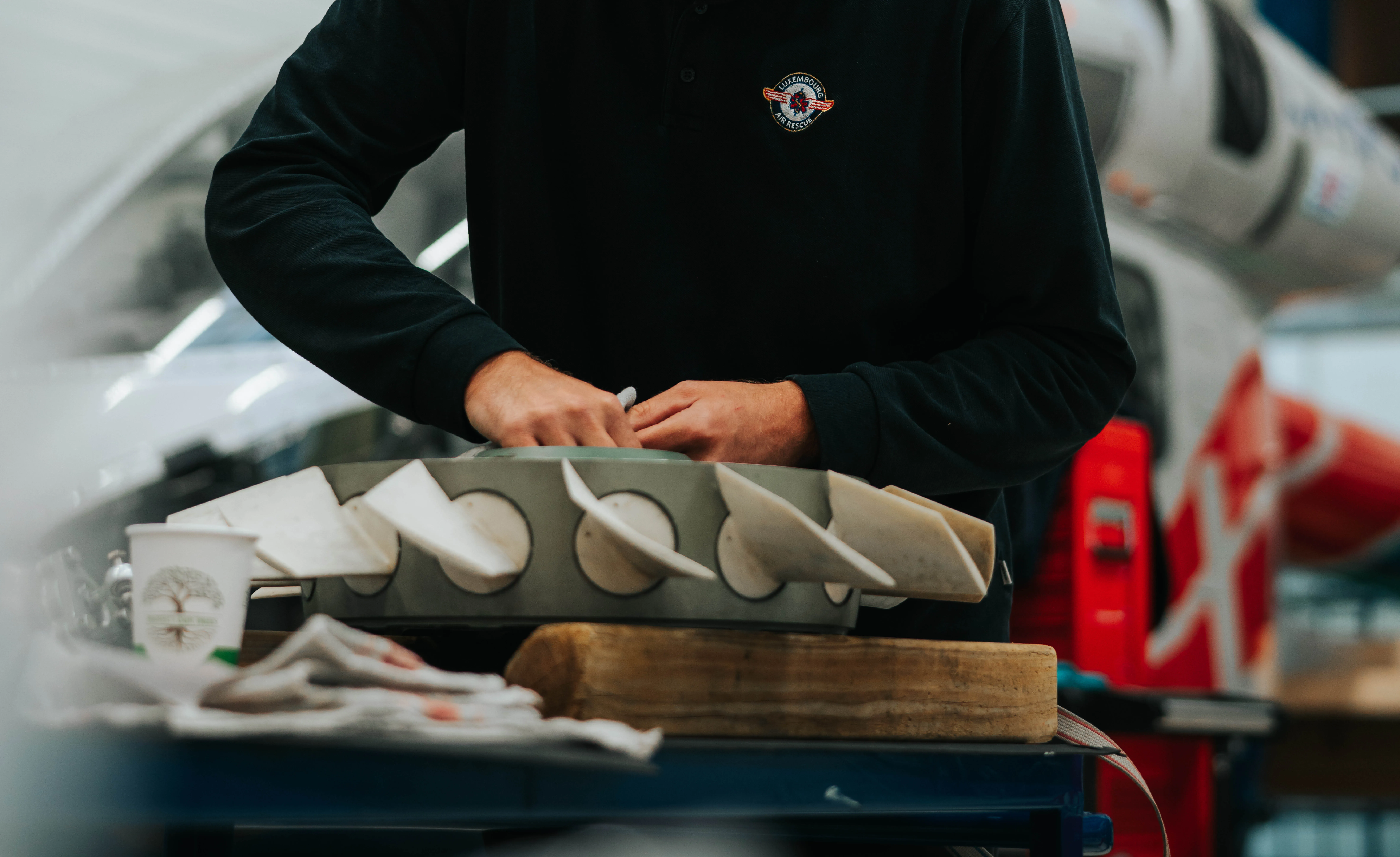 Personne portant une chemise Luxembourg Air Rescue travaillant avec un modèle aérodynamique gris avec des ailerons sur un bloc de bois.