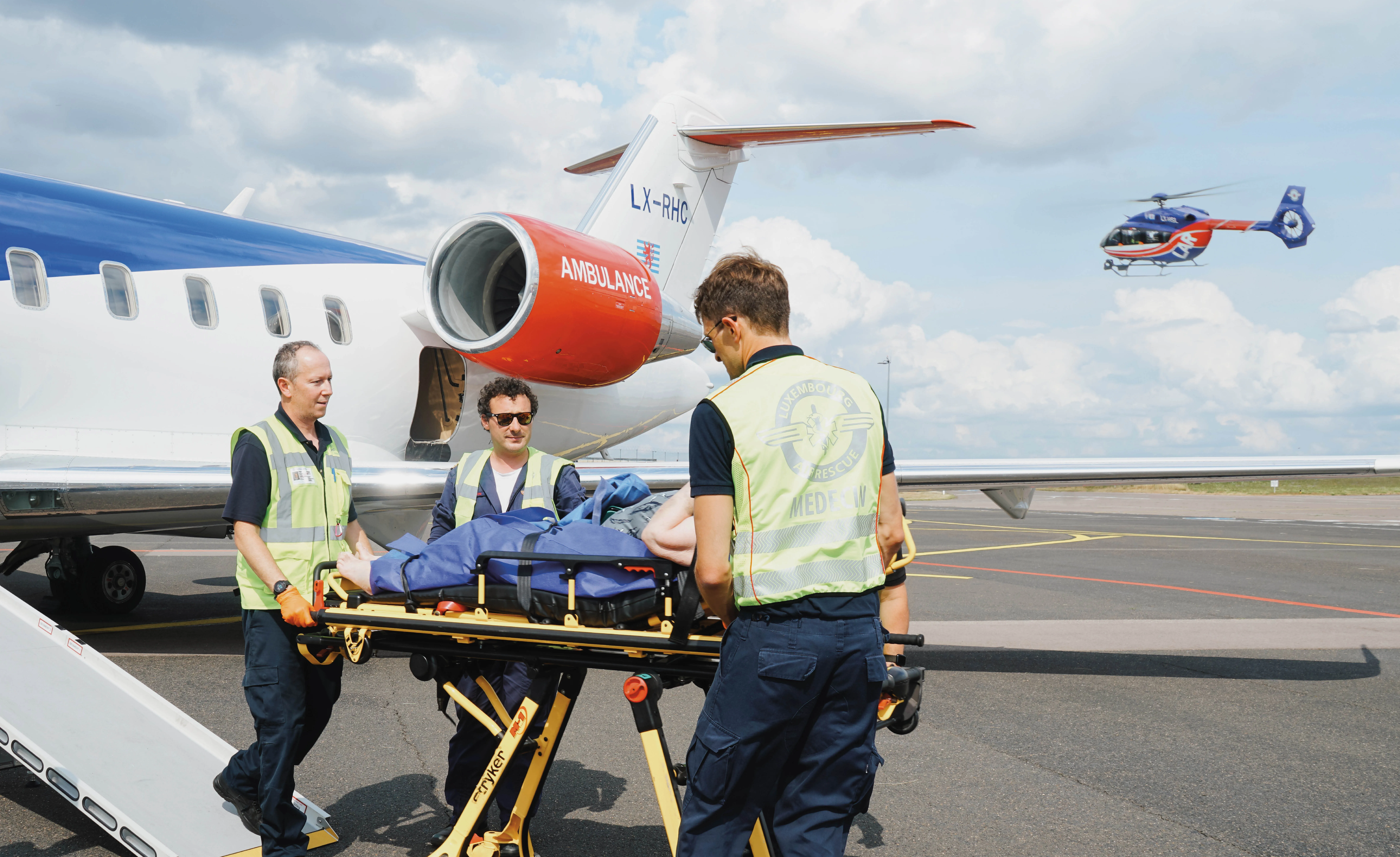 Une équipe médicale transfère un patient sur une civière à proximité d'un avion d'ambulance sur le tarmac d'un aéroport avec un hélicoptère médical volant en arrière-plan.