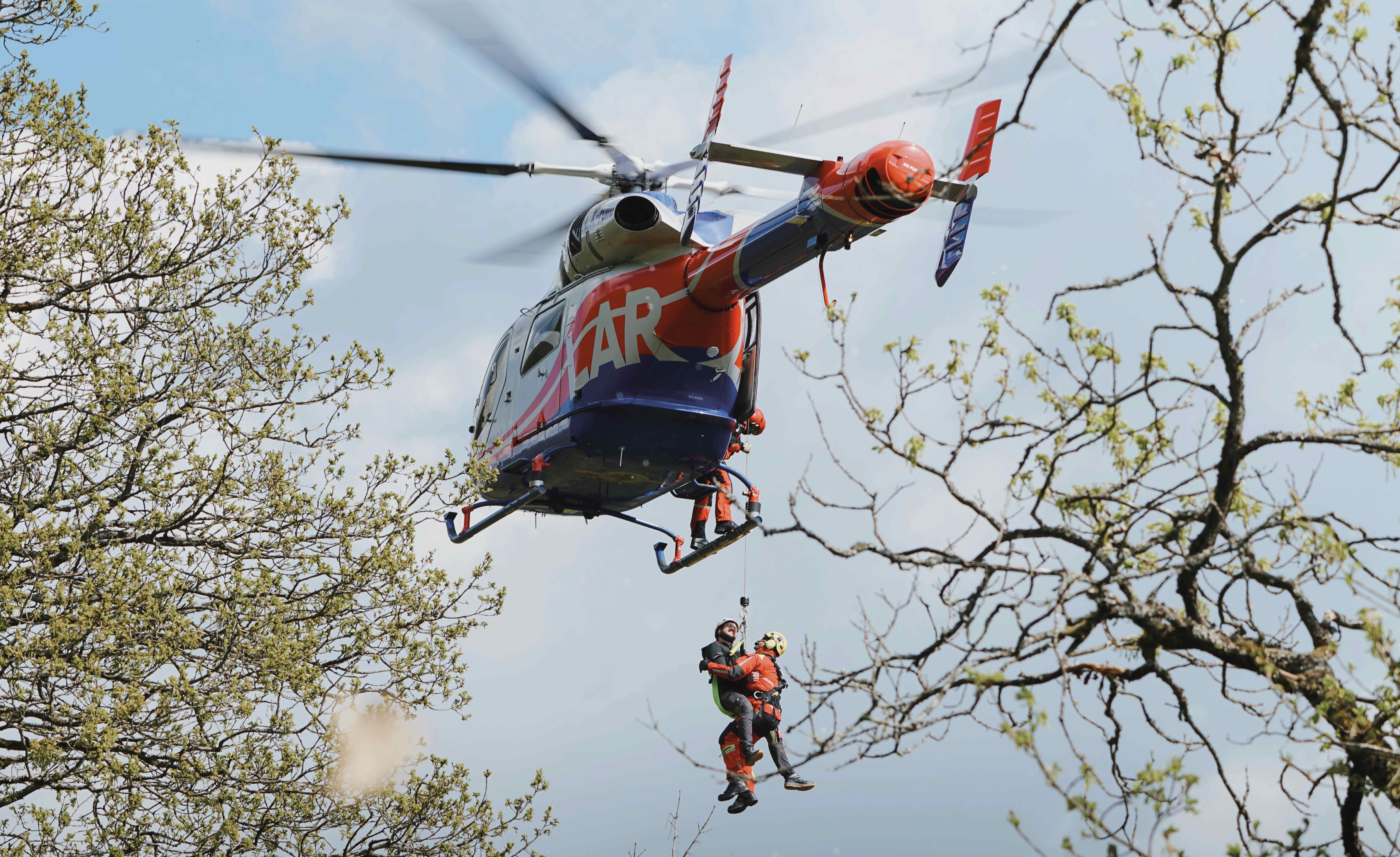 Hélicoptère de sauvetage transportant une personne à l'aide d'un harnais au milieu d'arbres avec un fond de ciel clair.