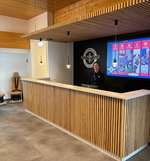 Reception area with a wooden slatted front desk, pendant lights, and a smiling receptionist standing behind the computer, with a Luxembourg Air Rescue logo and a digital training display in the background.