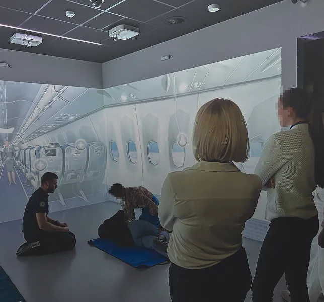 Group training session simulating a medical emergency inside an airplane cabin with a person lying on a mat and an instructor assisting.